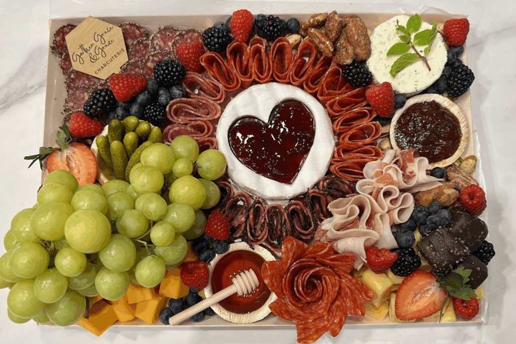 Charcuterie board with brie topped by a jam heart, salami rose, grapes, berries, cheese cubes, and honey dipper