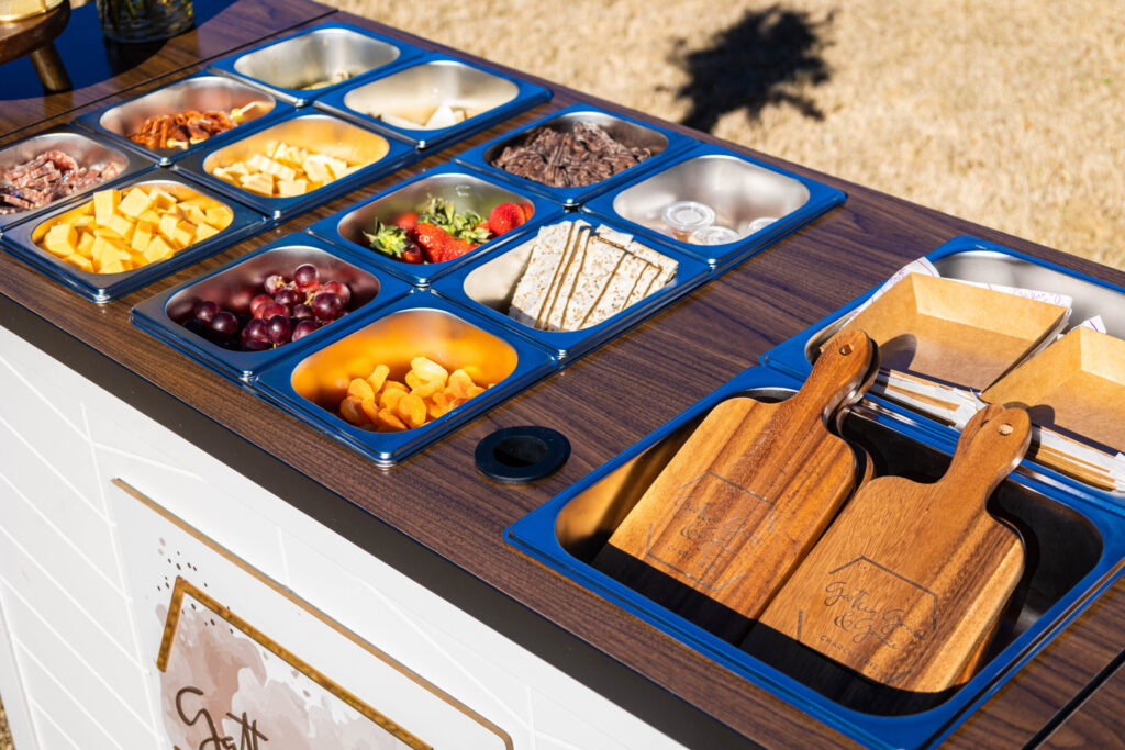 Mobile charcuterie bar with cheeses, cured meats, fruit, and crackers in blue trays beside wooden serving boards
